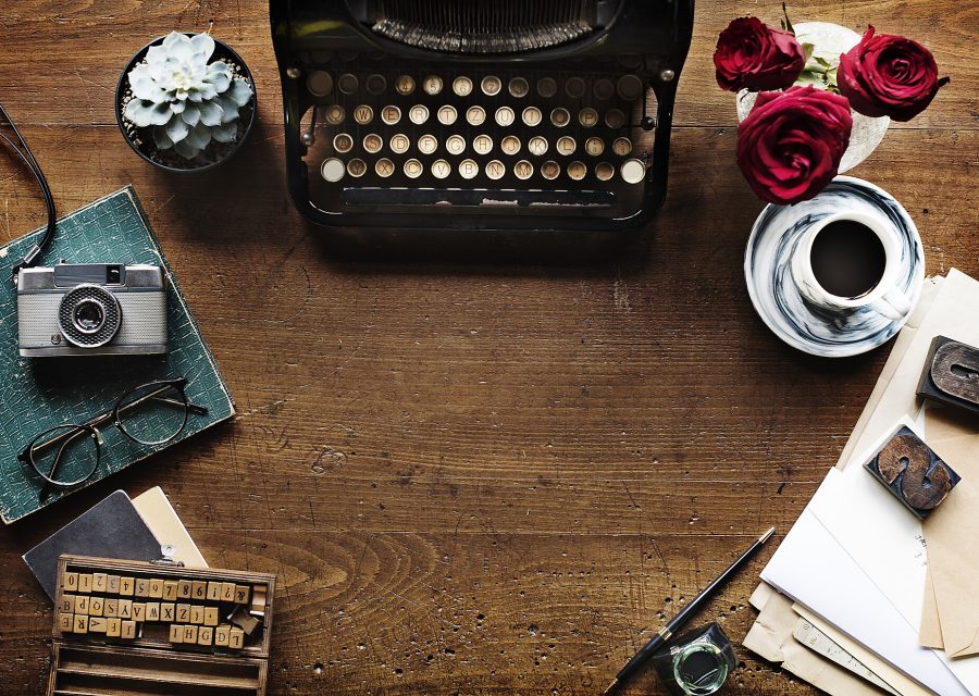 A flat-lay of a typewriter, cut flowers and a camera on a wooden desk.